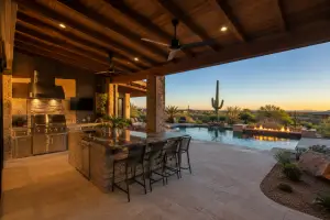 Covered outdoor living space in Phoenix Arizona with built-in kitchen area, stone patio, ceiling fans, pool, fire feature, and desert landscaping